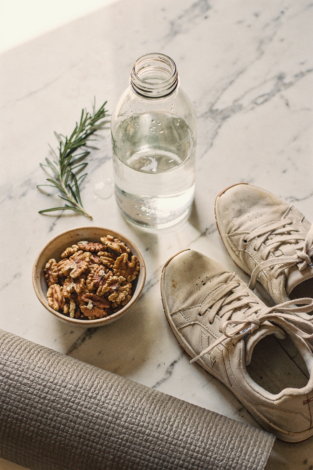 A glass water bottle half full, a small bowl of walnuts, a yoga mat corner rolled up, a sprig of rosemary, a pair of comfortable sneakers. , no faces, no hands
