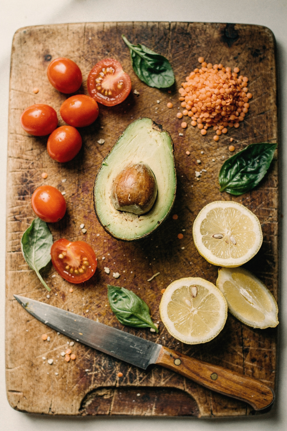 Halved avocado, cherry tomatoes, red lentils, lemon wedges, and basil on a wooden cutting board