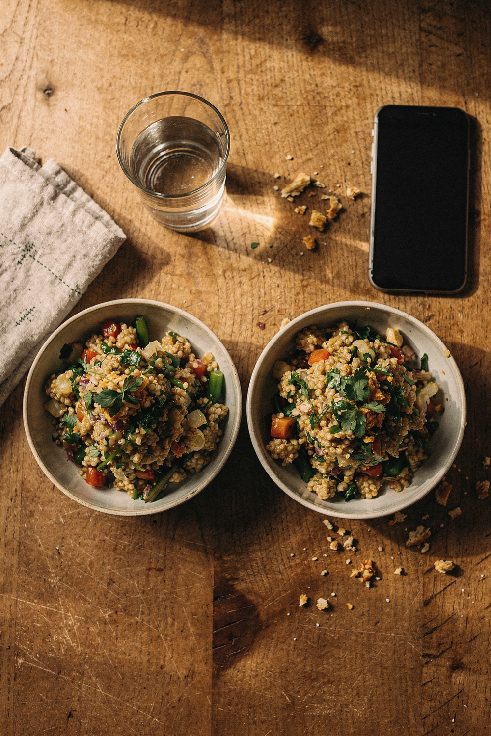 Two identical bowls of grain salad placed side by side, one with a napkin neatly placed and a glass of water, the other with a phone face-up and scattered crumbs. Contemplative, revealing, thoughtful. , no faces, no hands