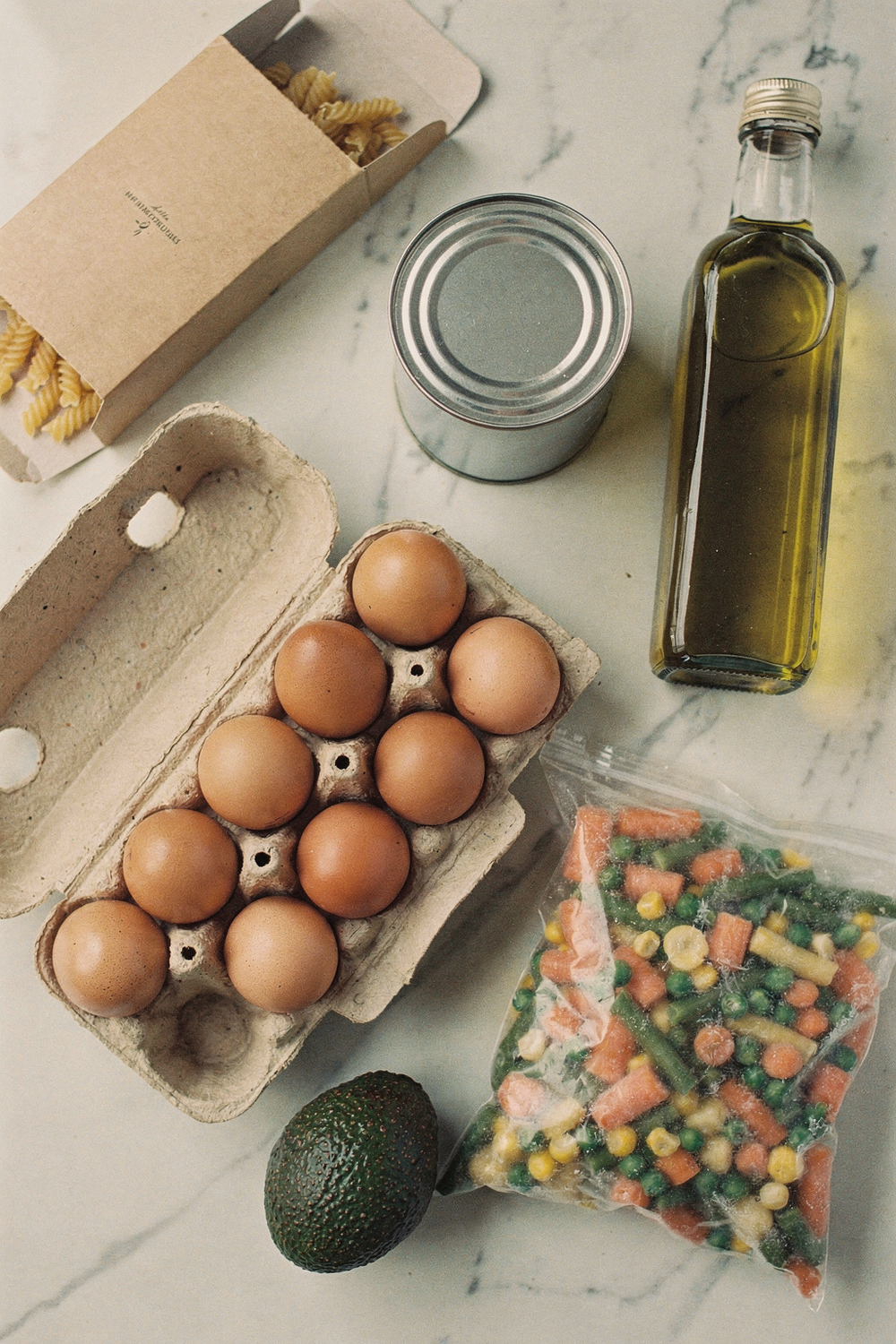A carton of eggs, a can of black beans, a bag of frozen vegetables, a box of pasta, a bottle of olive oil, an avocado. Approachable, practical, stocked-kitchen feel. , no faces, no hands