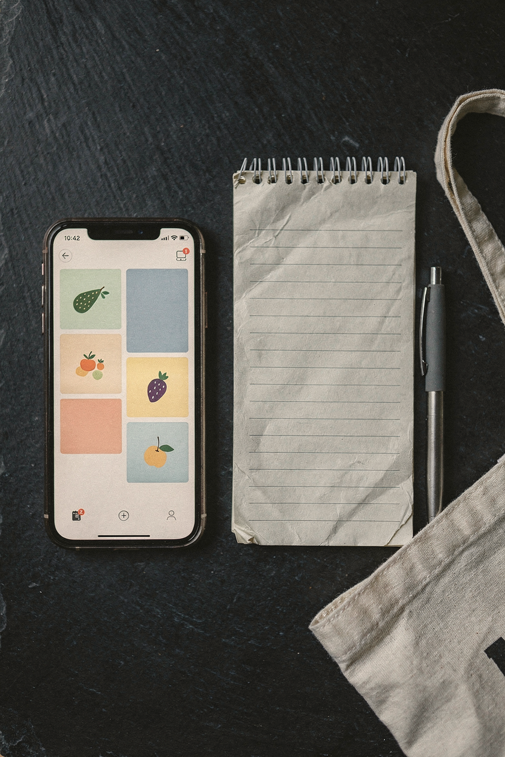 A phone showing a food delivery app next to a simple handwritten meal plan on a notepad, a grocery list, a pen, a reusable shopping bag folded flat. Honest, strategic, motivating. , no faces, no hands