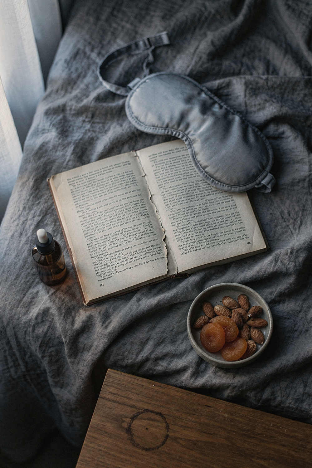 Sleep mask, open book, magnesium oil, and a bowl of almonds on rumpled grey bedsheets
