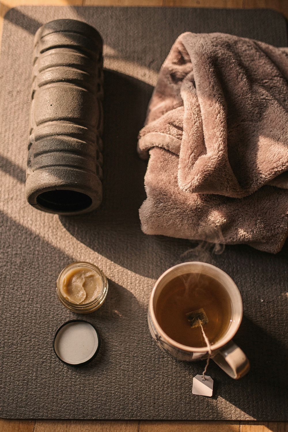 Foam roller, fleece blanket, muscle balm jar, and a mug of chamomile tea on a grey yoga mat