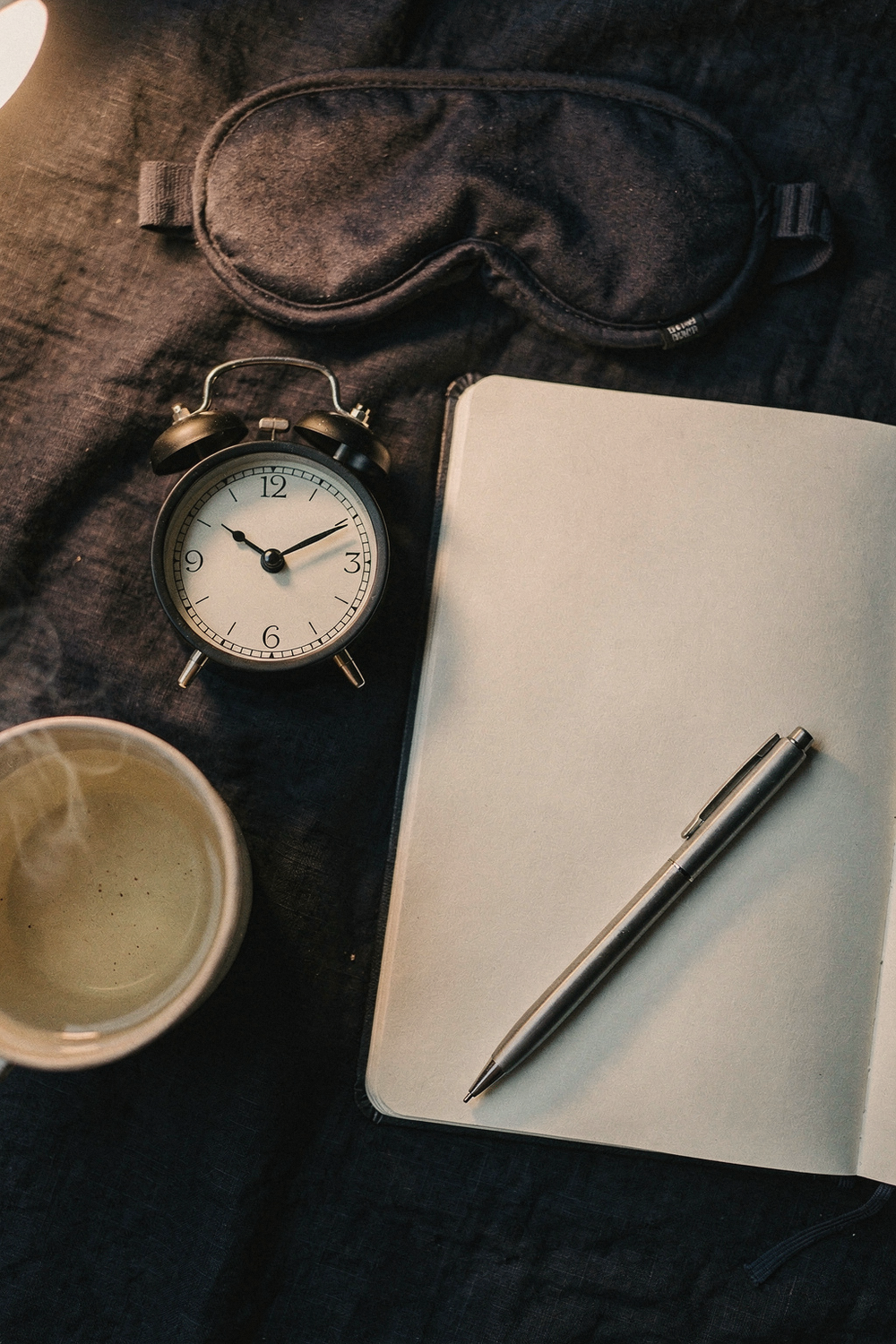 An alarm clock showing 2am, a journal open to a blank page, a pen, a cup of herbal tea, and a sleep mask. , no faces, no hands