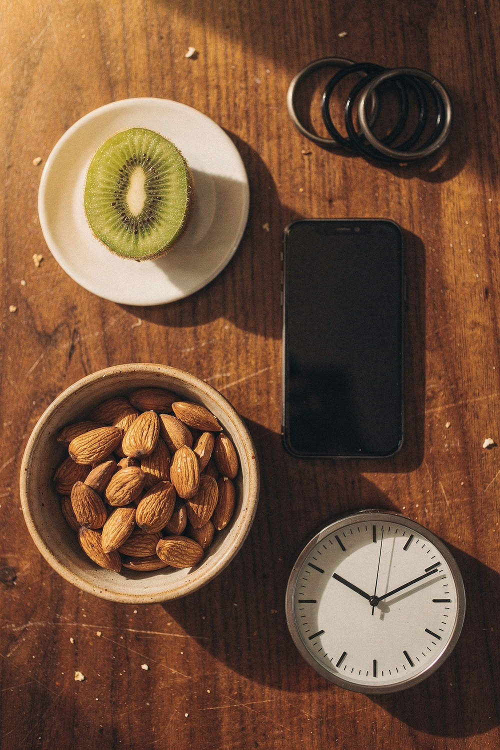 A bowl of almonds, a sliced kiwi on a small plate, a phone face-down, blackout curtain rings, and a simple analog clock. , no faces, no hands