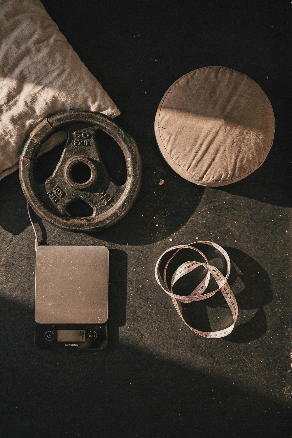A barbell plate, a kitchen scale, a pillow corner, a meditation cushion, and a body measurement tape arranged in two groups - one tight cluster, one spread apart