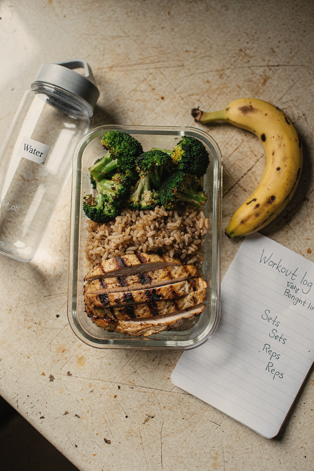 Meal prep container with sliced chicken, brown rice, and steamed broccoli beside a water bottle and banana