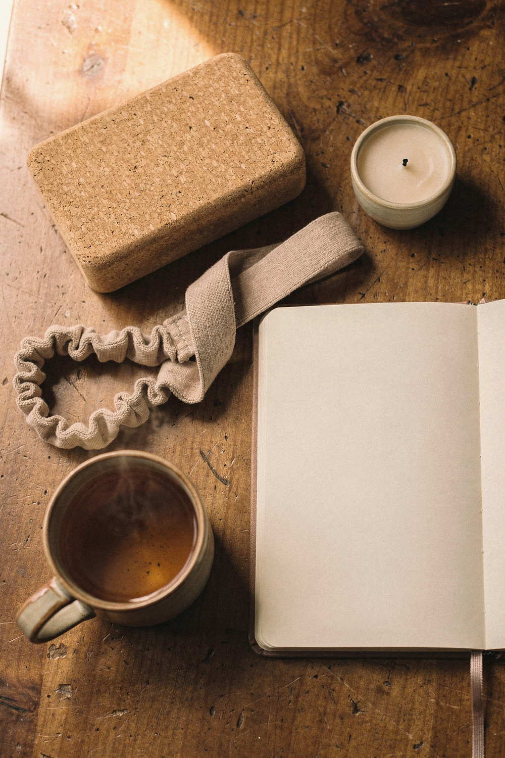 A cork yoga block, a journal open to a blank page, a herbal tea in a ceramic mug, a stretching strap, and a small candle unlit