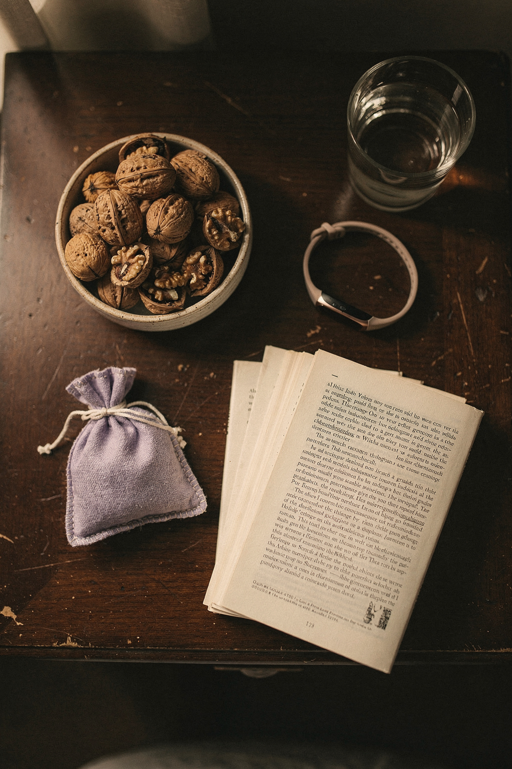 A bowl of walnuts, a sleep tracker, a lavender sachet, a small glass of water, and a book face-down. Restful, intimate