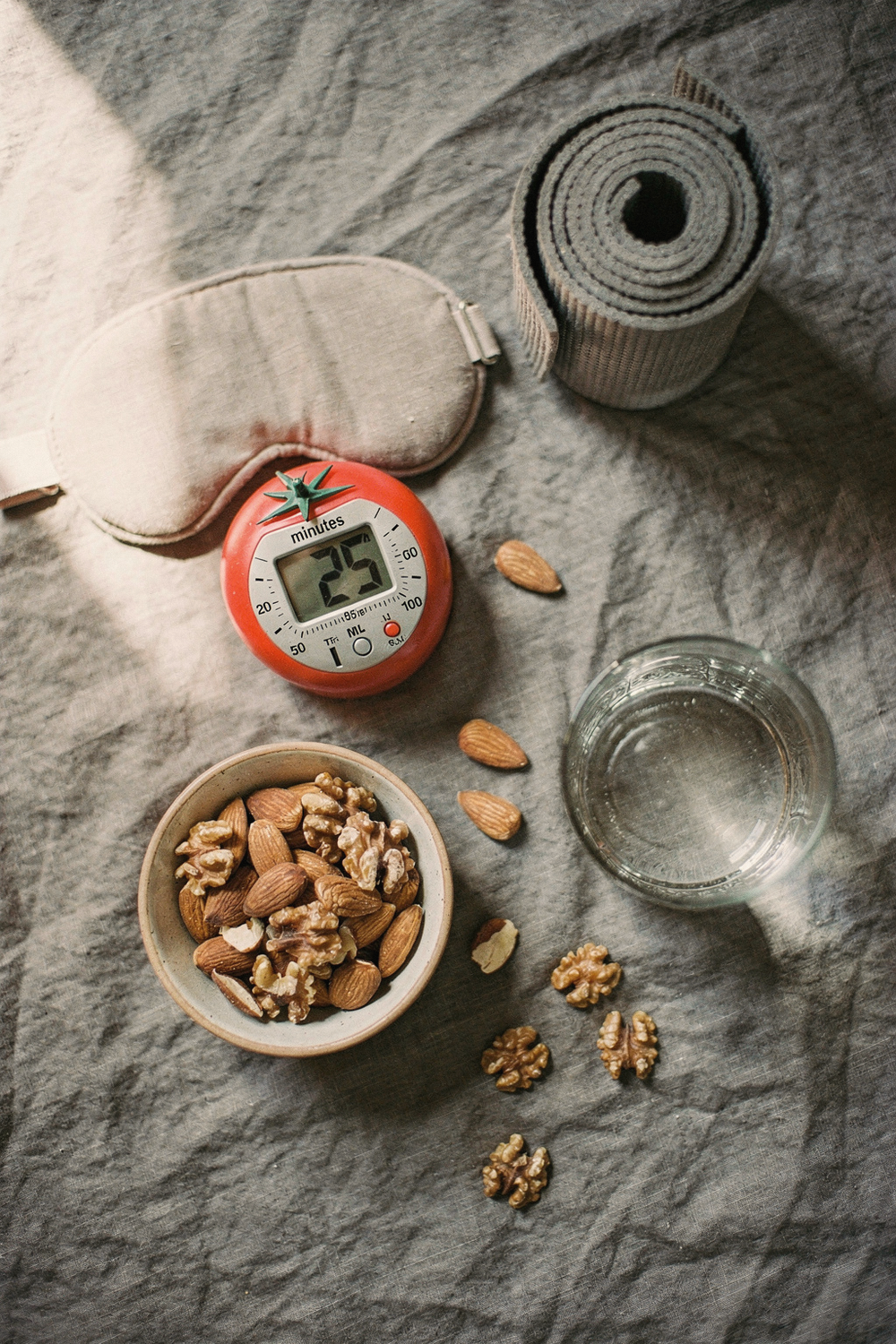 Pomodoro timer, sleep mask, a bowl of almonds, a rolled yoga mat, and a glass of water on a linen surface